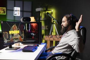 Cheerful gamer in apartment feeling relieved after finally receiving winner screen for killing all enemies. Joyous player congratulating himself after finishing videogame, sitting on gaming chair photo