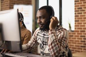 Closeup of a male telemarketer attentively listening to a client through wired headphones while looking at his computer screen. African American customer support agent seated at desk, using a headset. photo