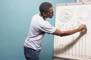 African american male professional in casual clothing drawing graphs and charts on a sketch board. Black man standing next to whiteboard and preparing data for presentation in blue background studio. photo
