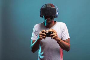 African American man wearing VR headset, holding a game controller and enjoying an immersive 3D gaming. Millennial guy facing the camera and playing a virtual reality game, against isolated backdrop. photo