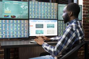 African american man working at trading desk at night, monitoring forex charts on multiple monitors. Black male employee working late in brick wall office, reviewing stock market growth in real time. photo