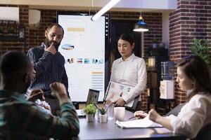 Business team reviews marketing strategies using digital charts on LED screen. Diverse colleagues leverage modern technology for financial analysis and online data insights during late night meeting. photo