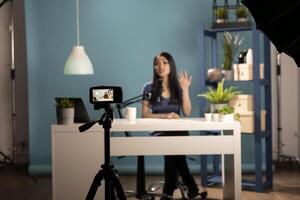 Social media influencer creating a review in a professional studio. Asian female blogger talking to the camera, seated at a desk, using a tripod, lighting equipment, and a blue backdrop. photo