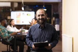Caucasian male entrepreneur holds a clipboard in modern startup office. The night shift setting, brick wall and blurred team in background emphasize productivity and teamwork. photo