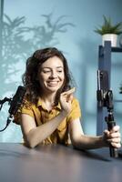 Jolly internet star talking in front on smartphone attached to selfie stick in studio, discussing with viewers. Happy woman using phone camera to record for social media photo