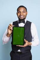 Smiling hotelier holding a tablet with isolated chroma key screen in studio, wearing a professional tuxedo uniform and greeting clients. Young hotel concierge against blue background. photo
