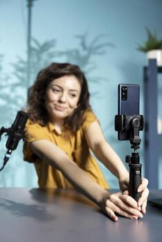Focus on smartphone attached to selfie stick used by joyous internet star to record in studio, discussing with viewers. Close up of phone used by cheerful woman using to record for social media photo