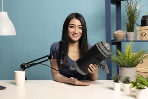 Cheerful female blogger holds up a strobe light in her studio, reviewing its features as she records social media content. She sits at desk with a microphone, engaging with her online audience. photo