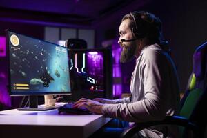 Man in neon lit apartment having fun with friends playing videogames, talking online using headphones mic, concentrating. Pro gamer using PC headset to join esports competition with teammates photo