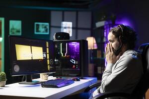 Player feeling sad after losing while playing multiplayer videogame, putting face in hands in frustration. Gamer at desktop PC feeling depressed after receiving game over screen photo
