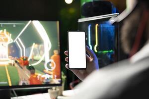 Man looking at mockup phone while playing SF FPS videogame, having fun in dark apartment room. Gamer holding isolated screen smartphone, competing in PvP internet multiplayer game, close up photo