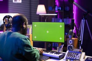 Musician operates on midi controller piano keys next to mockup screen, producing new songs with musical instruments and audio gear in home studio. Artist works with greenscreen. photo
