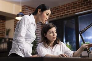 Dedicated female employees analyze research data on a laptop, having a business discussion. Asian manager stands and listens as her coworker explains the details, working together on a business task. photo