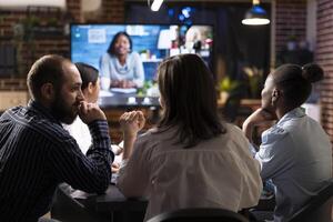 Business professionals collaborate in nighttime office meeting. A videoconference call is displayed on digital monitor as team members discuss marketing strategies and review financial data. photo