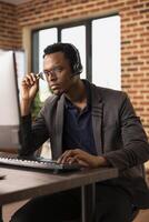 Pensive black man adjusting his glasses and working at desktop pc, focused on client information. Concentrated call center agent wearing headset, attending to customer support tasks on his computer. photo