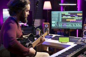 Young artist playing acoustic instrument accords in home studio with greenscreen layout shown on smartphone. Singer practices songs and tunes on guitar with strings, watching tutorials. photo