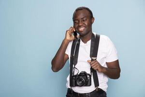African american man smiling brightly as he talks on his mobile phone, holding his suspenders with one hand. Cheerful photographer with a camera around his neck stands and speaks on the phone. photo