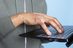 African american young guy typing on his laptop against blue background, composing emails and working on paperwork. Male overjoyed person multitasking in studio, cheerful adult. Close up. photo