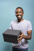Excited african american man carrying a black giftbox, delighted by the unboxing experience. Happy male individual holding a package, feeling thrilled about receiving a delivery. photo