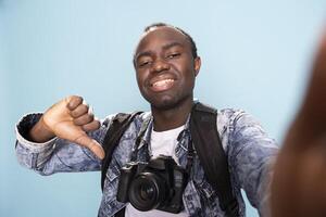 Happy african american photographer with a professional camera around his neck, giving thumbs down in studio. POV of a black male vlogger recording himself while making a playful disagreeing gesture. photo