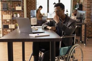 Determined employee with a physical injury, checking business analytics on his personal computer in modern workspace. Manager in wheelchair adjusting his glasses as he reviews data on his laptop. photo