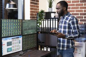 African american male investor standing with his tablet, monitoring forex charts on multiple screens. The office features a brick wall, creating a financial tech savvy workspace. photo