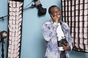 African american man grins widely while speaking on his mobile, holding his dslr device with one hand. Joyful male photographer with a camera around his neck stands, having a phone call in studio. photo