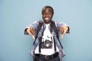 African American man in casual clothing, cheerfully giving two thumbs down while standing against isolated background. Smiling black male photographer with dslr camera, expresses disapproval in studio. photo