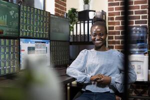 Black female entrepreneur smiling at the camera while preparing to review financial data across multiple monitors at her trading desk. The brick wall office adds a professional touch. photo