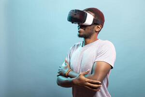 Young male individual enjoying an immersive VR experience, interacting with a 3D simulation. Portrait of african american tech enthusiast wearing virtual reality goggles, stands with crossed arms. photo