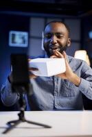 Happy tech content creator in dark studio filming technology review of newly launched digital device, unboxing it and presenting specifications to audience. Internet star showing product to viewers photo