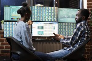 African american man and woman working late at night in office, analyzing stock market trends on multi screens. They collaborate at the desk, reviewing financial data and charts displayed on monitors. photo