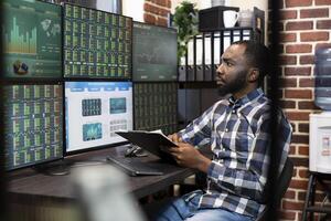 Male stock market trader with a clipboard in hand, analyzing real time data on a multi monitor setup in a dimly lit office. The scene reflects financial analysis and late night investment decisions. photo