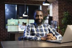 Portrait of african american man smiling and looking at the camera, seated at a desk with a laptop. The office is dimly lit with a brick wall, ideal for night shift work and stock market monitoring. photo