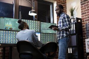 African American forex traders exchange investment strategies while tracking real time stock market transactions during night shift in brick wall office. Teamwork discussion at tech savvy workstation. photo