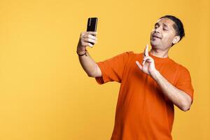 Young adult takes photos on his smartphone while he wears colorful clothes, showing a peace sign in front of the camera. Smiling indian person with man bum using his mobile phone in studio.
