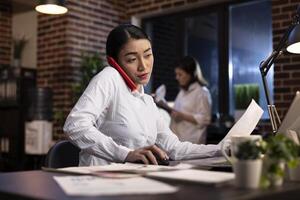 Busy woman juggles multiple tasks, having a phone call, holding a paper with one hand and mouse with the other, all while working on her laptop. Young woman managing late night work responsibilities. photo
