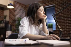 Caucasian female manager working tirelessly at her desk, leaning on one hand while scrolling through her laptop. A fatigued business analyst reviews company research data with an exhausted expression. photo