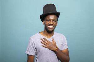 Young gentleman wearing a top hat and with his hand placed on his chest, standing against an isolated background. Portrait of african american guy with a black headpiece, smiling at camera. photo