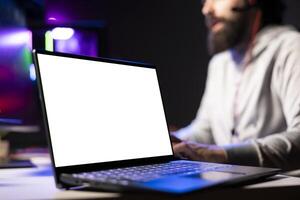 Person in dimly lit apartment playing games on isolated screen laptop at computer desk, enjoying day off from work. Gamer battling enemies in online multiplayer shooter on mockup notebook device photo