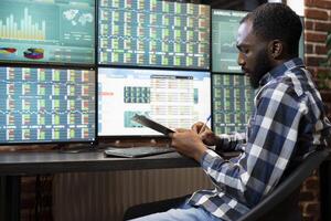 African American manager sitting and writing on clipboard, recording progress updates for company hedge fund. Male professional taking notes, working on the risk analysis of stock trading in office. photo