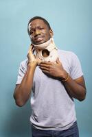 Hurt male individual using a neck collar in a studio setting to recover from a fractured vertebra, visibly upset from pain. Young black man standing in discomfort with his hands on the cervical brace. photo
