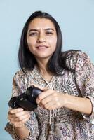 Relaxed indian person entertained by games on modern console, enjoying leisure activity against blue background. Female player enjoying game on joystick, having fun defeating enemies. photo
