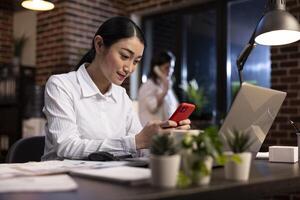 Female employee taking a break at work, using her mobile device at her office desk during her night shift. Young asian manager texting on her smartphone, communicating with remote colleagues. photo
