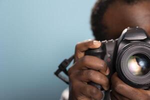 Closeup of black male photographer using a professional camera in a studio with a blue background. Selective focus on an african american man holding and demonstrating how to use a DSLR. photo