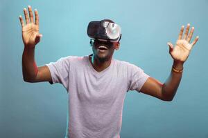 Millennial man raising hands and playing a futuristic game, enjoying immersive 3D simulation. African american person with an amazed expression, using VR glasses against a blue background. photo