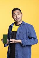 Indian content creator presenting green screen on mobile device, standing against orange background in the studio. Young male model wearing casual clothes and a man bun, internet channel. photo