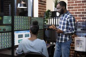 African american team of two stockbrokers working late, reviewing forex charts and financial data on multiple monitors in brick wall office. Male manager with a tablet collaborates with his coworker. photo