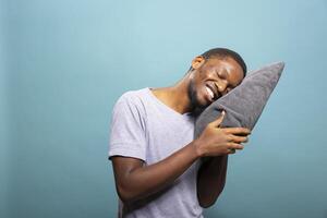 Young black man standing and leaning on a gray cushion with a bright smile, excited for a small nap. Cheerful african american person with eyes closed, resting his head on a pillow. photo