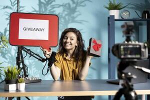 Happy influencer announcing giveaway for viewers with cardboard sign, offering gift box, engaging with audience. Joyous woman organizing contest, explaining rules using copy space board photo